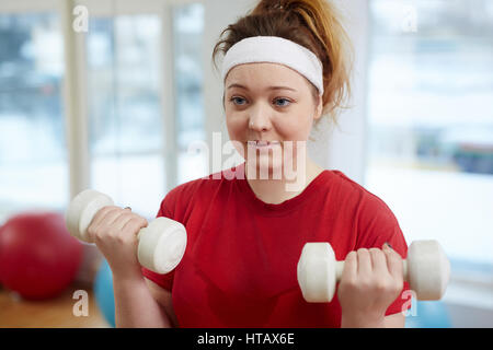 Portrait of cute overweight woman working out in fitness studio:  doing weightlifting exercise with dumbbells Stock Photo