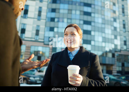 Portrait of young smiling businesswoman chatting with African-American colleague outside office building and drinking coffee from disposable cup Stock Photo