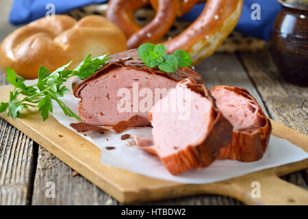 Traditional oven fresh Bavarian meat loaf with a brown crispy crust on a wooden cutting board garnished with parsley and oregano, a roll and a pretzel Stock Photo