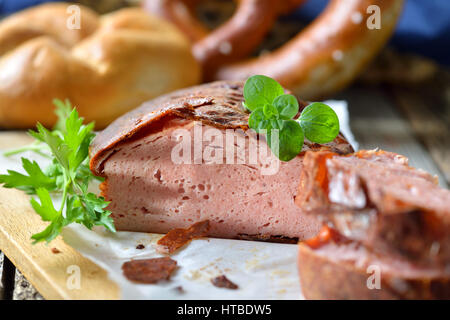 Traditional oven fresh Bavarian meat loaf with a brown crispy crust on a wooden cutting board garnished with parsley and oregano, a roll and a pretzel Stock Photo