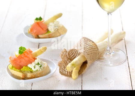 Delicious canapes with white asparagus, cream cheese with herbs, smoked salmon on Italian ciabatta bread with lettuce, served with a glass of wine Stock Photo