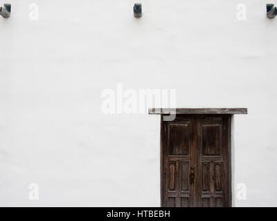 A rustic dark brown wooden door stands out against the whitewashed wall of an adobe building. Stock Photo