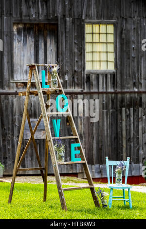Wooden ladder on grassy lawn with "L O V E" letters placed on steps with old rustic wooden barn in the background and old wooden painted chair Stock Photo