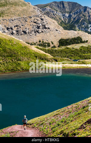 Female hiker along hillside mountain trail leading down toward colourful alpine mountain lake with mountains in the background Stock Photo