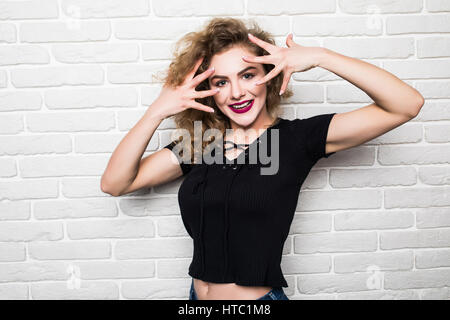 Closeup portrait of happy, excited successful young woman giving peace, victory or two sign, isolated white bricks background. Positive emotions, face Stock Photo