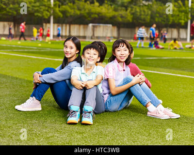 group of happy smiling elementary school boys and girls sitting on grass of playground. Stock Photo