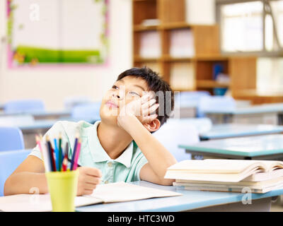 asian elementary schoolboy thinking while doing homework in classroom, looking up hand on cheek. Stock Photo