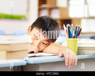 asian elementary schoolboy looking tired and exhausted resting his head on desk in classroom. Stock Photo