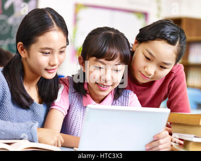three asian elementary school girls friends looking at a tablet together during break in classroom. Stock Photo