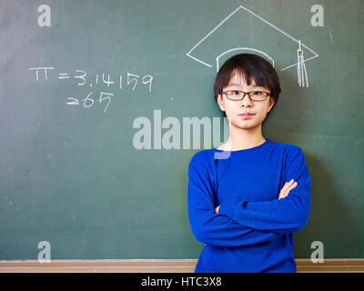asian elementary school boy wearing glasses standing under a doctoral hat drawn with chalk on blackboard. Stock Photo