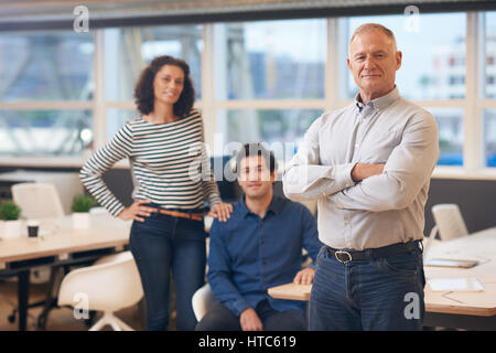 Portrait of a mature businessman standing confidently with his arms crossed in a modern office with two young work colleagues in the background Stock Photo