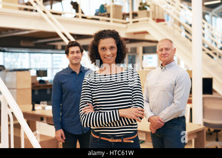 Portrait of a smiling businesswoman standing confidently with her arms crossed in a modern office with two male colleagues in the background Stock Photo