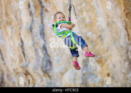 Little girl in climbing gear hanging on rope attached to safety harness Stock Photo