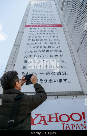 A man takes pictures of a huge banner on display outside Sony building in Ginza on March 11, 2017, Tokyo, Japan. The banner displayed by Yahoo Japan Corp. is marked with a red line at 16.7 meters, the actual height of tsunami which hit the northeast coast of Japan on March 11, 2011. 2017 marks the sixth anniversary of the earthquake and tsunami that killed thousands and led to the Fukushima nuclear crisis. Credit: Rodrigo Reyes Marin/AFLO/Alamy Live News Stock Photo