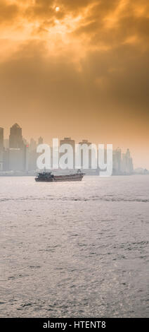 A Chinese Cargo Ship in the Victoria Harbour with the skyscrapers and the Hong Kong skyline, with the sun shining through dramatic clouds. China. Stock Photo