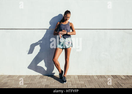 Full length shot of fitness woman standing against a wall checking fitness progress on her smart watch. Fit female runner relaxing after workout outdo Stock Photo