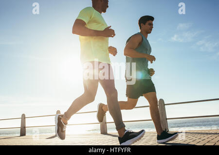 Low angle shot of two young men running on sea front. Fit young runner working out on a sunny day. Stock Photo