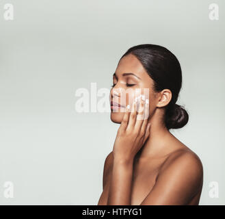 Beautiful young woman with eyes closed while applying moisturizer to her clean face. Close up portrait female model applying face cream against grey b Stock Photo