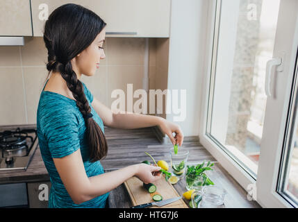 happy young woman making detox water with cucumber, mint and lemon at home kitchen. Healthy drink. Weight loss dieting concept. Process of preparing e Stock Photo