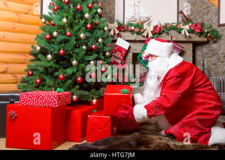Bearded Santa Claus puts gift boxes under christmas tree, fireplace on background Stock Photo