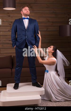 Beautiful bride in white dress and veil makes marriage offer to the groom in suit standing on table, wooden room interior on background Stock Photo