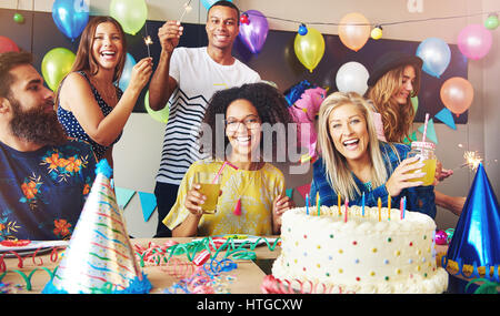 Six young adults celebrating a birthday party while holding drinks as they sit around a table with cake and party hats Stock Photo