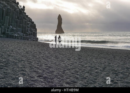 Moody view silhouettes of couple on black lava beach with sea stack and basalt cliff face, Reynisfjara, Iceland in winter Stock Photo
