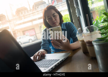 Young Asian woman using smartphone while working in coffee shop. Freelance worker on workday with laptop concept Stock Photo