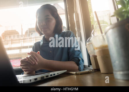 Young Asian woman looking at her laptop computer while working in coffee shop. Freelance worker with online business activity concept. Hipster in digi Stock Photo