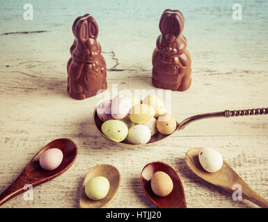 Easter sugar coated candy eggs on wooden and silver spoons with chocolate bunnies on white wooden background Stock Photo