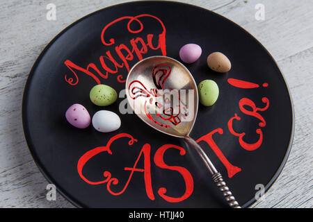 Happy Easter hand lettering quote closeup written on black plate with Easter bunny drawing on vintage silver spoon. Decorated with chocolate easter eg Stock Photo