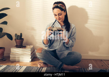 Pretty trendy young woman checking a photo on the back of her camera as she relaxes barefoot on the living room floor in the sunlight from a window Stock Photo