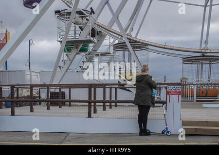 Barry Island, south Wales, 12th March 2017. A single woman stands by the entrance to the Ferris Wheel ride in Barry Island Pleasure Park. The sky is dark and overcast, and a childs scooter is resting against the wall. Barry Island was full of people making the most of the dry weather today Credit: Gary Parker/Alamy Live News Stock Photo