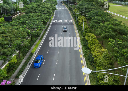 Roadway with four lanes and cars on them in Singapore. On the sides there are street lamps and a lot of green trees. It is an exterior view from the M Stock Photo