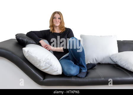 Grinning long haired young woman is sitting on a black and white couch with black and white pillows. Isolated on the white background. Woman is lookin Stock Photo