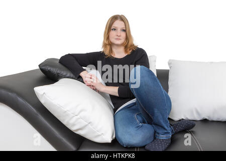 Pensive long haired young woman is sitting on a black and white couch with black and white pillows. Isolated on the white background. Stock Photo