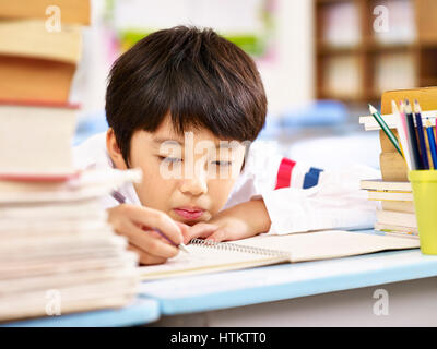 tired and bored asian elementary school boy doing homework in classroom, head resting on desk. Stock Photo