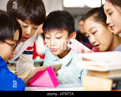 group of asian elementary school children gathering around playing game together using tablet during break. Stock Photo