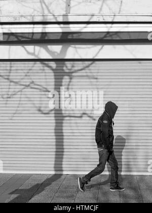 Young man wearing a hoodie walking past closed shop shutters with the shadow of a tree.  Street photography in Plymouth Devon England UK Stock Photo