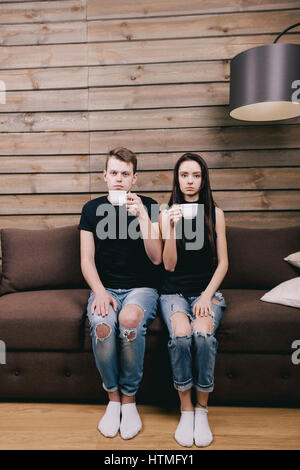 Cute couple sitting on a brown couch, sadly looking into the camera and drinking hot tea. concept of a first date or modest people Stock Photo