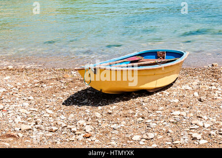 A dinghy painted yellow outside and blue inside rests on a stony shingle beach with its oars shipped close to the blue clear seawater of Lulworth Cove Stock Photo