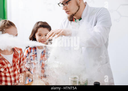 Interaction of chemical reagents. Serious nice experienced scientist performing an experiment and being surrounded by smoke while having an optional c Stock Photo