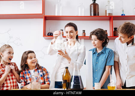 Look at it. Beautiful cheerful female scientist standing among the children and holding a flask while being in a good mood Stock Photo