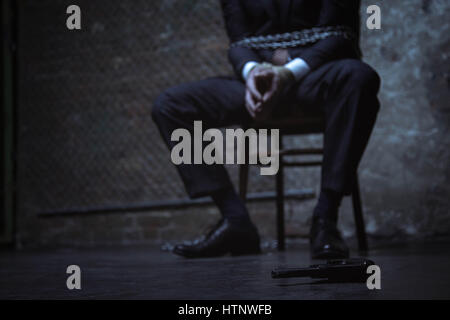 How can I get near it. Classy typical rich businessman being tied up and sitting on a chair while hoping getting to a gun lying close to him Stock Photo