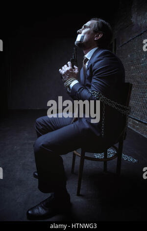 I have a chance now. Scared desperate captured gentleman sitting in a dark room while being tied up to a chair with chains and holding a gun in his ha Stock Photo