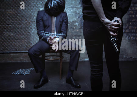 What are we gonna do next. Protectless unable captured executive sitting tied to a chair with chains while his kidnapper staring at him with a gun in  Stock Photo