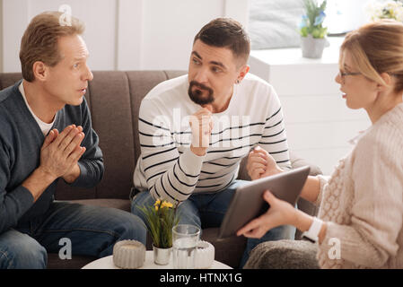 Psychological help. Unhappy worried nice man looking at the psychologist and speaking with her while sitting on the sofa with his boyfriend Stock Photo