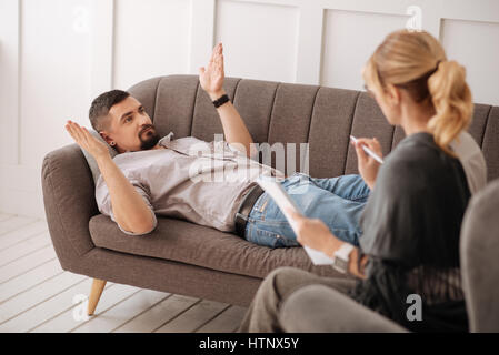 What should I do. Handsome nice bearded man lying on the sofa and asking for advice while visiting a psychologist Stock Photo
