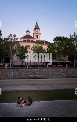 Three young girls back to camera sitting by the River Genil across from the Hermandad de Los Escolapios as dusk shows the moon in a clear sky Stock Photo