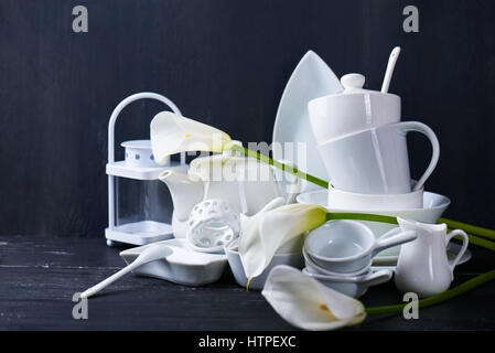 Set of new white dishes with teapot, tea cups, and plates on a black wooden table with fresh callas Stock Photo